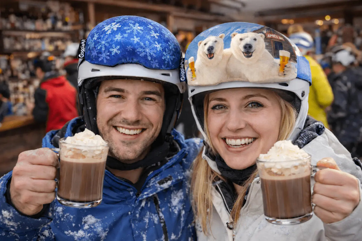 Two people wearing ski goggle covers holding hot chocolate mugs in a snowy setting